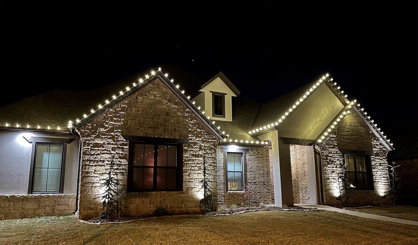 A brick home outlined in warm white Christmas roofline lights at night