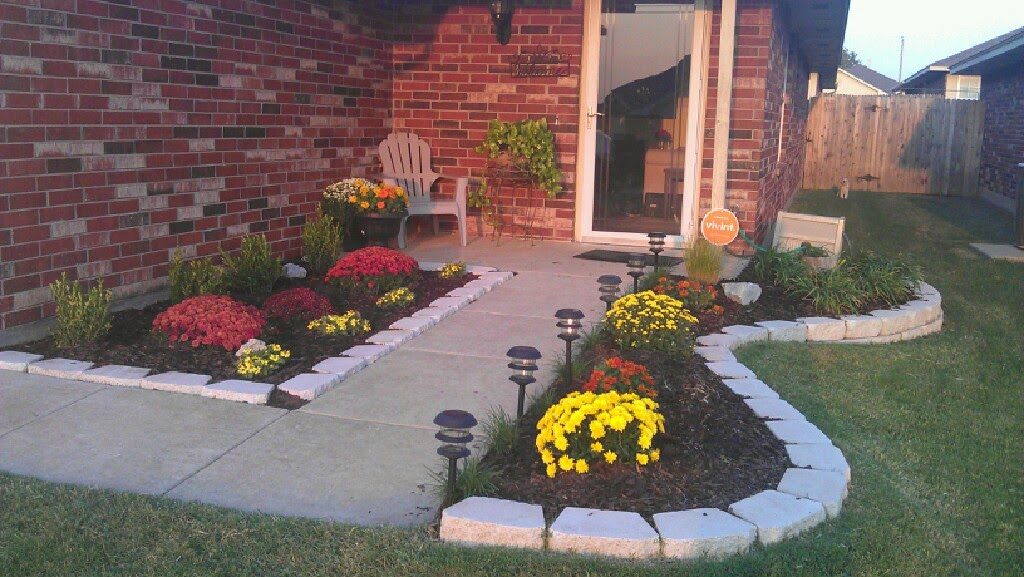 Tidy paver-edged flower bed with red and yellow mums in front of a brick home